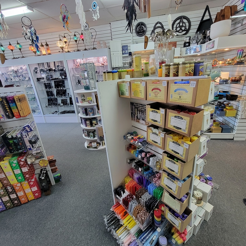 Back centre view of The Zen Shop Oshawa showcasing a vibrant display of spiritual candles, including votives, tall spell candles, chime candles, and holders, with shelves of incense and tarot cards nearby, and crystal jewellery cabinets in the background.
