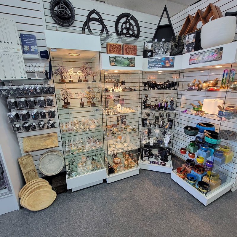 Back south corner of The Zen Shop Oshawa featuring crystal wire-wrapped jewellery, gemstone trees, witchcraft tools such as cauldrons, chalices, incense holders, and crystal balls, as well as rare crystals like Moldavite and meteorites. Also shown are Tibetan singing bowls, copper products, and decorative wooden plaques.